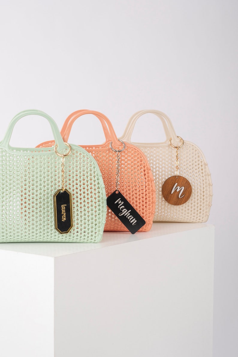 Pu&ograve; includere: Tre borse a rete colorate con etichette in legno personalizzate. Le borse sono verde chiaro, pesca e bianco. Le etichette riportano "Lauren", "Meghan" e "M".