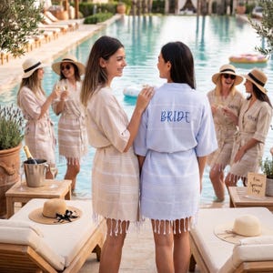K&ouml;nnte beinhalten: Mehrere Frauen in hellen Badem&auml;nteln mit Fransen stehen in der N&auml;he eines Pools. Ein Bademantel ist hellblau mit dem Wort "BRIDE" in blauer Schrift auf dem R&uuml;cken. Andere Badem&auml;ntel sind beige mit wei&szlig;en Streifen. Einige tragen Strohh&uuml;te.