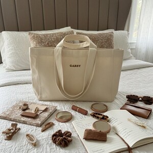 Op de afbeelding: Een beige canvas draagtas met de naam "GABBY" geborduurd op de voorkant, omringd door accessoires zoals haarclips, scrunchies, een notitieboek, lippenstift en zonnebril. De items zijn gerangschikt op een wit sprei.