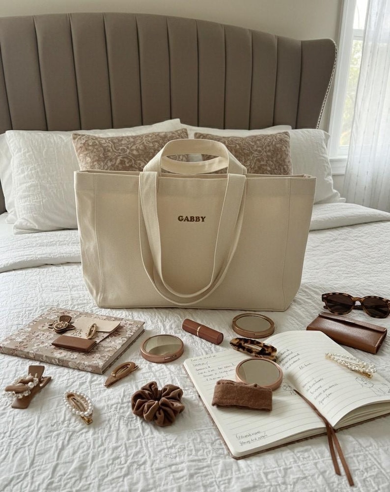 Op de afbeelding: Een beige canvas draagtas met de naam "GABBY" geborduurd op de voorkant, omringd door accessoires zoals haarclips, scrunchies, een notitieboek, lippenstift en zonnebril. De items zijn gerangschikt op een wit sprei.