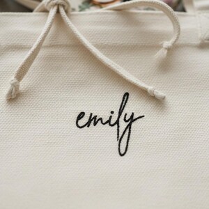 Op de afbeelding: Cr&egrave;mekleurige canvas draagtas met een trekkoordsluiting. Het woord "emily" is in zwart script op de voorkant geborduurd. Het trekkoord van de tas is gemaakt van een dik, gebroken wit touw.