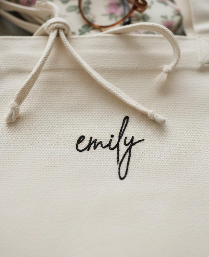 Op de afbeelding: Cr&egrave;mekleurige canvas draagtas met een trekkoordsluiting. Het woord "emily" is in zwart script op de voorkant geborduurd. Het trekkoord van de tas is gemaakt van een dik, gebroken wit touw.