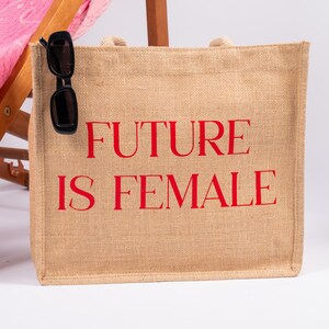 May include: A beige jute tote bag with red text that reads "FUTURE IS FEMALE". A pair of black sunglasses is hanging from the top of the bag.