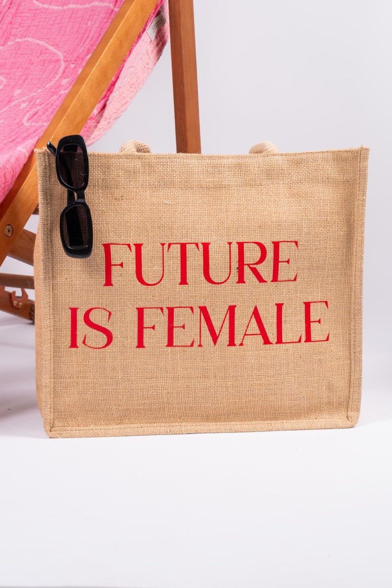 May include: A beige jute tote bag with red text that reads "FUTURE IS FEMALE". A pair of black sunglasses is hanging from the top of the bag.