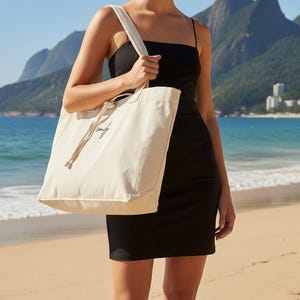 May include: A large, off-white canvas tote bag with two shoulder straps and a knotted drawstring closure. The bag features a small, black cursive script logo on the front. The material appears to be a sturdy, woven fabric with a natural, unbleached finish.