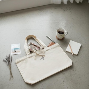 May include: A cream-colored canvas tote bag featuring the name emily in black cursive script on the front. The bag has two top handles and a drawstring closure. Inside the bag are floral patterned glasses and a small brown leather wallet.