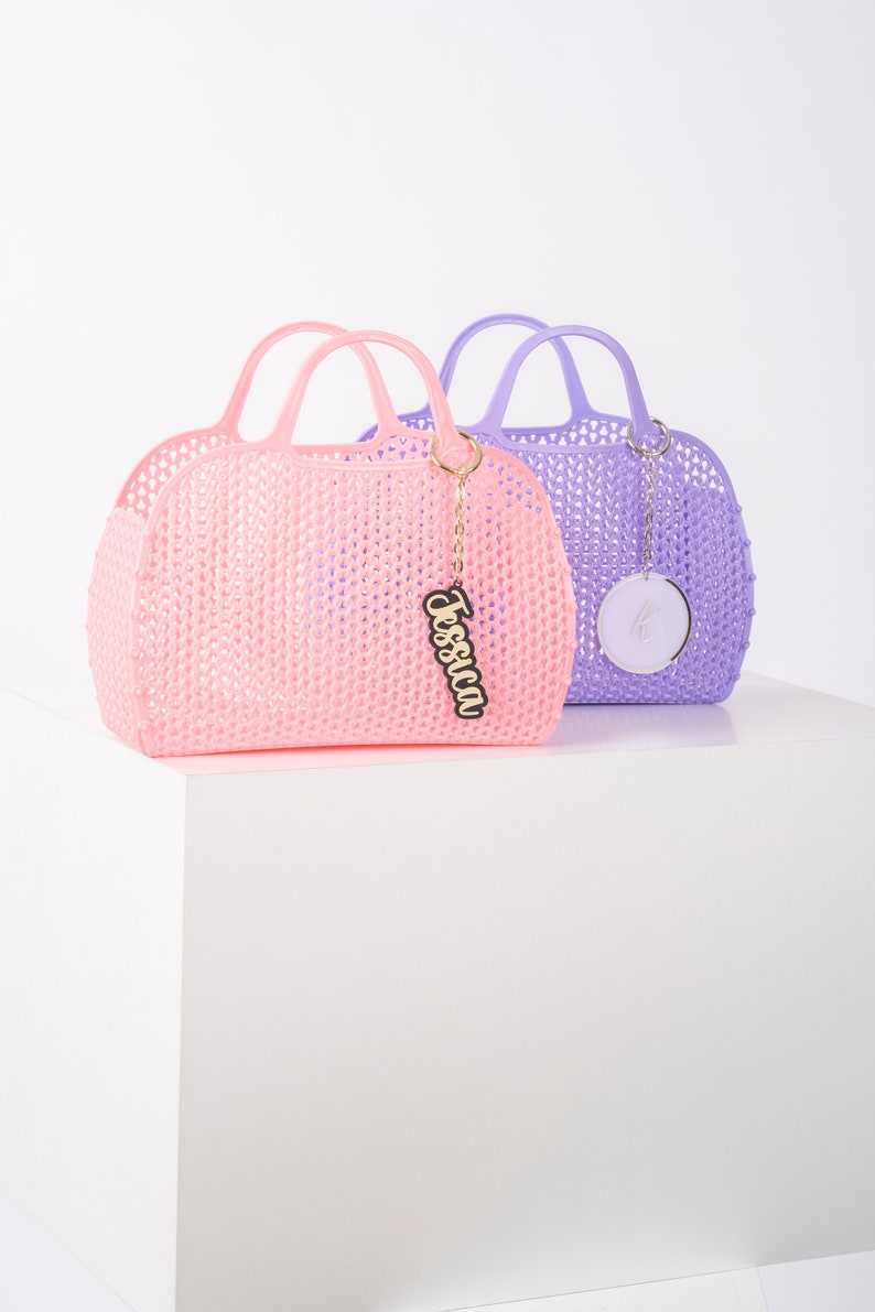 Pu&ograve; includere: Due borse a mano in plastica con manici. La borsa a sinistra &egrave; rosa e ha un ciondolo dorato con il nome "Jessica" su di esso. La borsa a destra &egrave; viola e ha un ciondolo rotondo argentato.