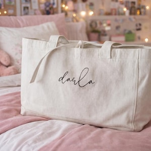 May include: A rectangular off-white canvas tote bag featuring two sturdy top handles and the name Darla embroidered in black cursive script on the front center. The bag has a structured base and a clean, minimalist design with visible fabric texture.