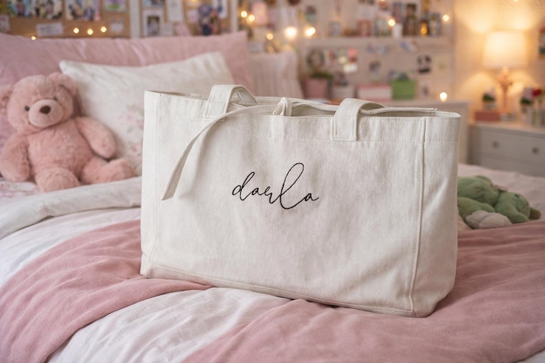May include: A rectangular off-white canvas tote bag featuring two sturdy top handles and the name Darla embroidered in black cursive script on the front center. The bag has a structured base and a clean, minimalist design with visible fabric texture.