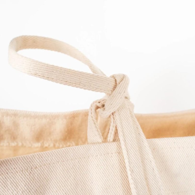 Op de afbeelding: Close-up van een beige canvas draagtas. De handgreep van de tas is bovenaan geknoopt, met een lus die omhoog loopt. De stof van de tas heeft een textuur en de achtergrond is effen wit.