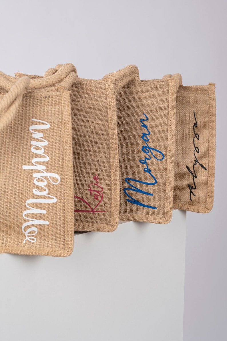 May include: Four burlap tote bags with rope handles. The bags are lined up in a row and each has a name embroidered on the front in different colors:  Meghan in white, Katie in red, Morgan in blue, and Alyssa in black.