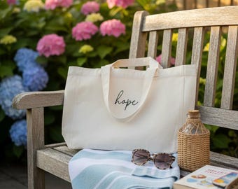 May include: A cream-colored tote bag with the word "hope" in script, resting on a wooden bench. The scene includes a striped blue and white towel, sunglasses, a decorative bottle, and a book. Colorful hydrangeas bloom in the background.