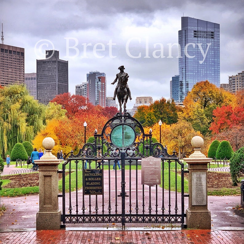 May include: A black wrought iron gate with a sign that reads "Bicycling & Roller Blading Prohibited" in front of a park with fall foliage and a statue of a man on horseback. The statue is in front of a tall building in the background.