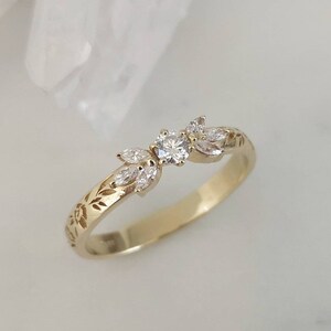 May include: A delicate gold band ring with a floral design and a small round diamond in the center. The ring is set with several smaller diamonds in a leaf shape on either side of the center stone.