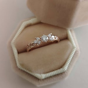 May include: A delicate rose gold ring with a floral design featuring a round centre stone and smaller accent stones. The ring is displayed in a velvet ring box.