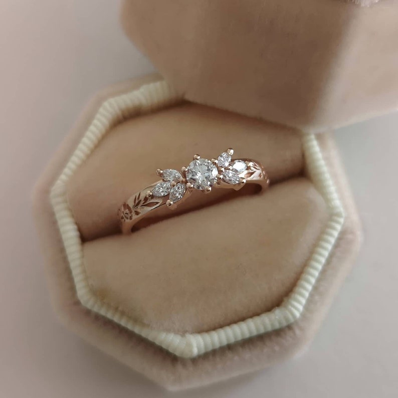 May include: A delicate rose gold ring with a floral design featuring a round centre stone and smaller accent stones. The ring is displayed in a velvet ring box.