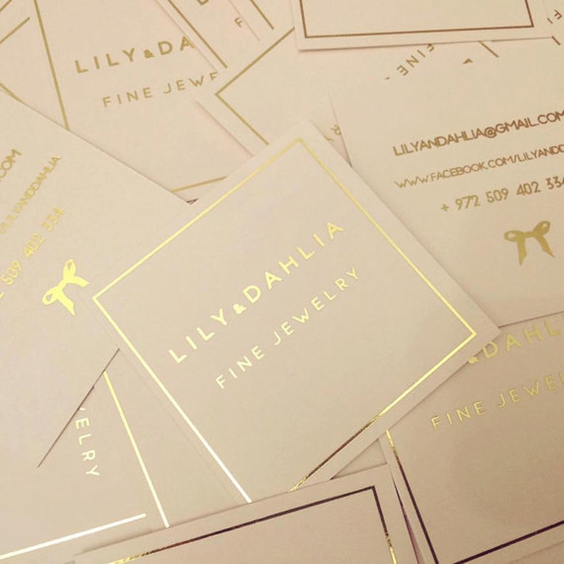 Op de afbeelding: Close-up van verschillende ivoren visitekaartjes met gouden letters. De tekst op de kaartjes luidt "Lily & Dahlia Fine Jewelry" en bevat contactgegevens.