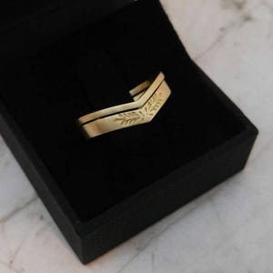 May include: A pair of gold wedding bands with a leaf design. The bands are displayed in a black velvet box.