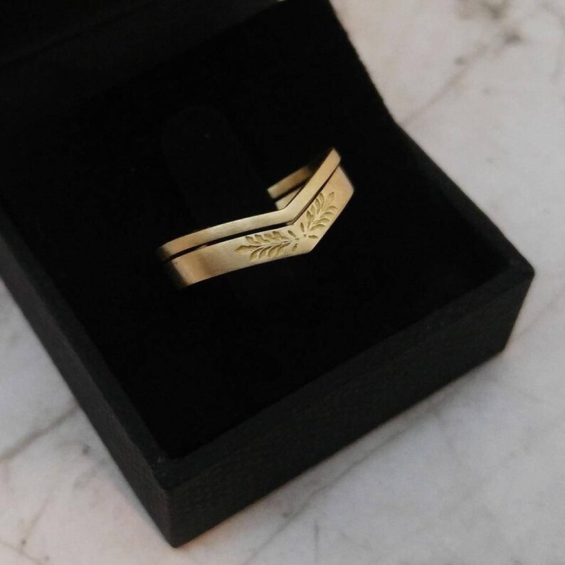 May include: A pair of gold wedding bands with a leaf design. The bands are displayed in a black velvet box.