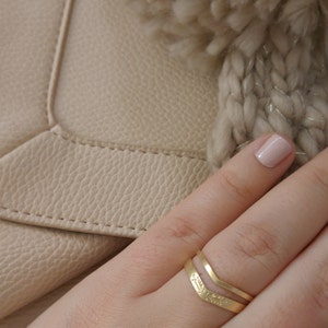 May include: A gold ring with a chevron design and floral details. The ring is worn on a finger with a light pink nail polish.