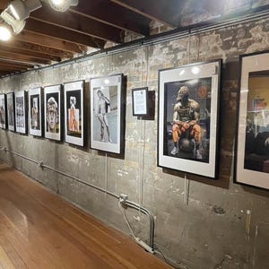 May include: An art gallery featuring framed photographs and artwork. The gallery has a wooden floor and exposed brick walls. Several framed images are displayed along the wall, showcasing various artistic styles and subjects. The lighting is provided by track lighting.