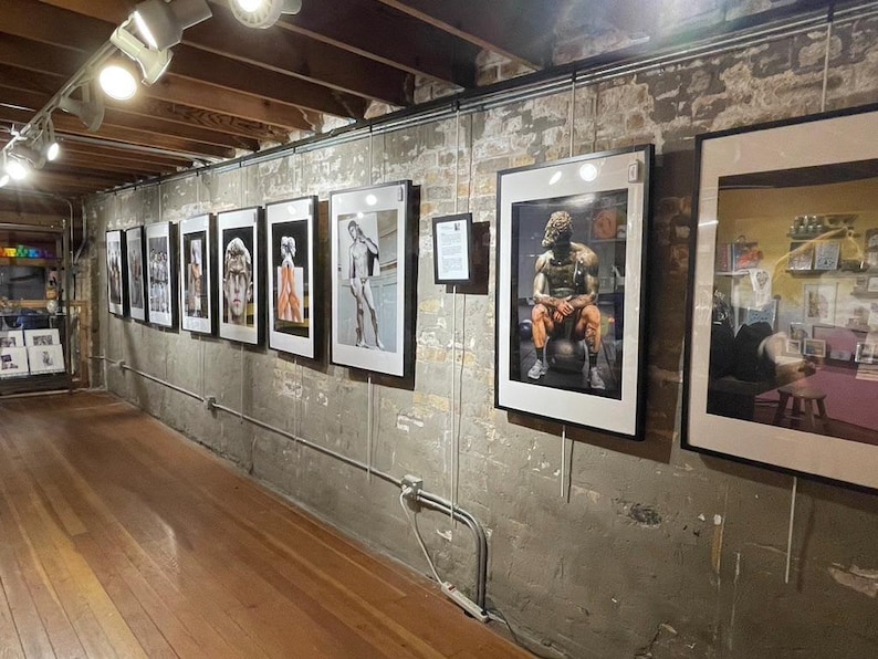 May include: An art gallery featuring framed photographs and artwork. The gallery has a wooden floor and exposed brick walls. Several framed images are displayed along the wall, showcasing various artistic styles and subjects. The lighting is provided by track lighting.