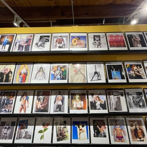 May include: A display of framed art prints featuring various artistic styles and subjects. The prints are arranged on shelves, showcasing a diverse collection of images, including portraits, figures, and abstract compositions. The frames are black, and the art is displayed in a gallery setting.