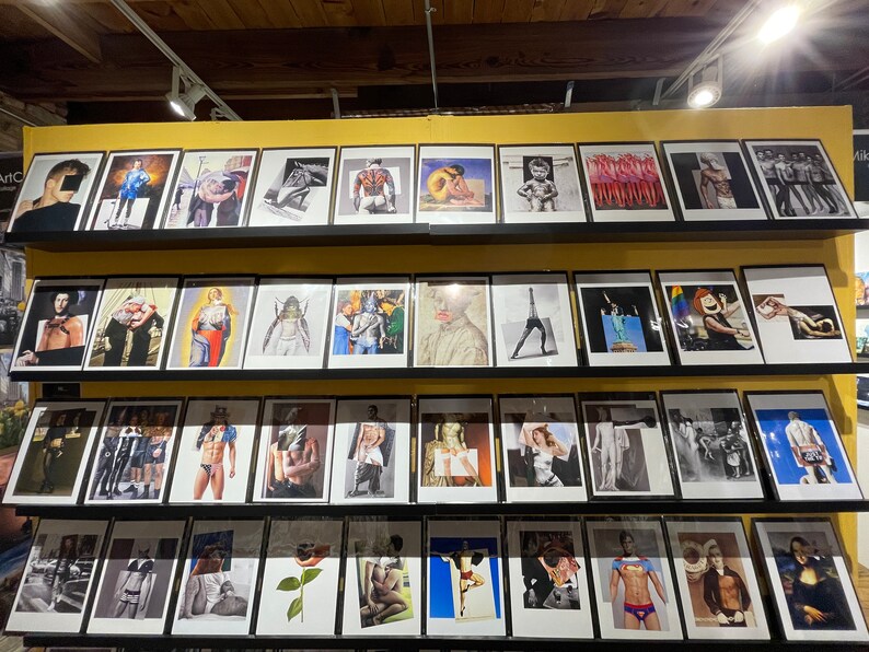 May include: A display of framed art prints featuring various artistic styles and subjects. The prints are arranged on shelves, showcasing a diverse collection of images, including portraits, figures, and abstract compositions. The frames are black, and the art is displayed in a gallery setting.