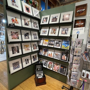 May include: A retail display featuring framed art prints and greeting cards. The prints showcase various images, including portraits, abstract art, and pop culture references. The display includes a sign that says "NEW! Arrivals".