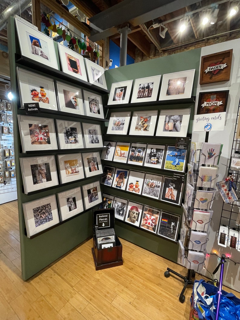May include: A retail display featuring framed art prints and greeting cards. The prints showcase various images, including portraits, abstract art, and pop culture references. The display includes a sign that says "NEW! Arrivals".