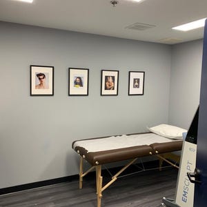 May include: A medical examination room with a massage table, framed artwork, and a door with a "CHICAGO" sign. The room has gray walls, dark flooring, and a white countertop with supplies.