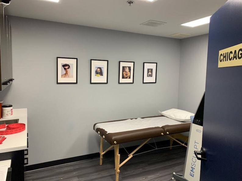May include: A medical examination room with a massage table, framed artwork, and a door with a "CHICAGO" sign. The room has gray walls, dark flooring, and a white countertop with supplies.