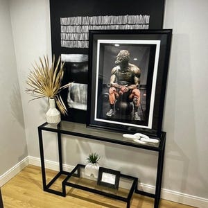 May include: A modern black metal console table with a glass top, displaying framed art and decorative items. The art includes a black and white abstract piece and a framed photograph of a statue. A white vase with dried palm fronds and a small plant are also on display.