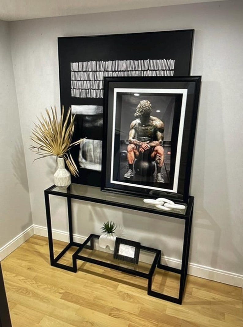 May include: A modern black metal console table with a glass top, displaying framed art and decorative items. The art includes a black and white abstract piece and a framed photograph of a statue. A white vase with dried palm fronds and a small plant are also on display.