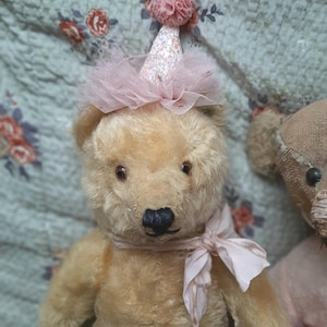 Puede incluir: Un oso de peluche vintage con pelaje marrón claro, que lleva un gorro de fiesta rosa y una bufanda a juego. El oso tiene ojos oscuros y una nariz negra. El fondo es una tela con estampado floral.