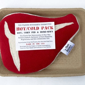 May include: A red and white plush steak-shaped hot/cold pack, labeled "The Fleece Butchery's Therapeutic Hot/Cold Pack." The pack is made of fleece and eco-felt, and is filled with corn. It rests in a brown cardboard tray.