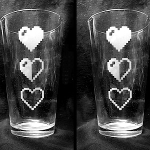 May include: Two clear glass pint glasses with three white pixelated hearts etched on the front of each glass.