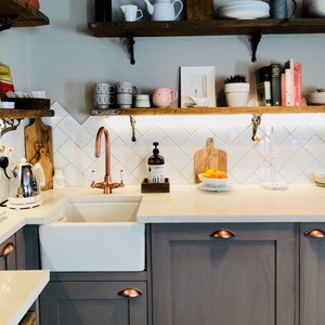 May include: A white farmhouse kitchen sink with a copper faucet, a wooden cutting board, and a bowl of oranges. The kitchen has white subway tile backsplash and dark gray cabinets with copper handles.