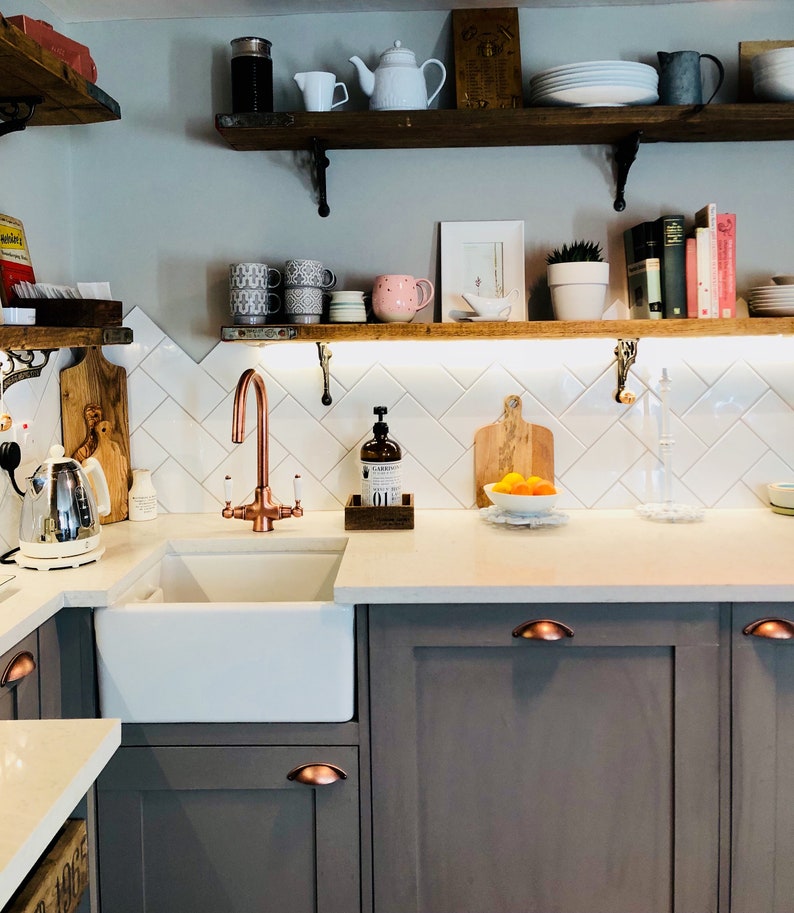 May include: A white farmhouse kitchen sink with a copper faucet, a wooden cutting board, and a bowl of oranges. The kitchen has white subway tile backsplash and dark gray cabinets with copper handles.