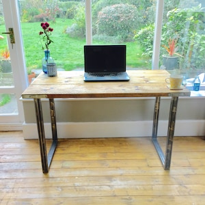 May include: A rustic wooden desk with metal legs. The desk has a natural wood finish and is supported by two black metal legs. A laptop computer is on the desk, along with a cup of coffee and a bottle of water.