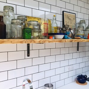 May include: A wooden shelf with metal brackets is mounted on a white tiled wall. The shelf is filled with various glass jars, bottles, and other items. There is a framed print on the wall above the shelf.