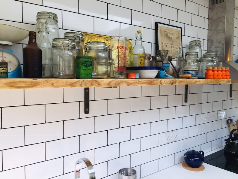 May include: A wooden shelf with metal brackets is mounted on a white tiled wall. The shelf is filled with various glass jars, bottles, and other items. There is a framed print on the wall above the shelf.