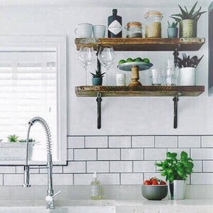 May include: Two wooden shelves with metal brackets, mounted on a white tiled wall. The shelves are filled with various items, including glass bottles, plants, and cups. A silver faucet is visible in the foreground.