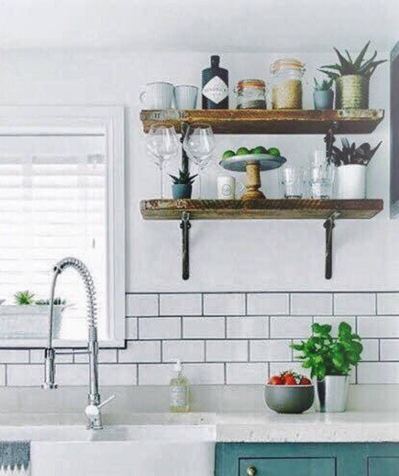 May include: Two wooden shelves with metal brackets, mounted on a white tiled wall. The shelves are filled with various items, including glass bottles, plants, and cups. A silver faucet is visible in the foreground.