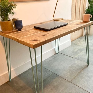 May include: A wooden desk with a natural finish and a light wood grain. The desk has four hairpin legs painted a light blue color. A laptop computer is on the desk, along with a potted plant and a mug.