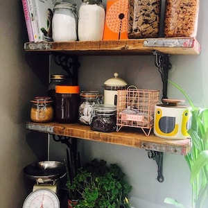 May include: A rustic wooden shelf unit with three shelves, filled with various jars and containers. The top shelf has two glass jars with orange lids, a glass jar with a white lid, and a book titled "Baking". The middle shelf has a variety of jars with different contents, including sugar, tea, and coffee. The bottom shelf has a vintage kitchen scale, a potted plant, and a blue pitcher.
