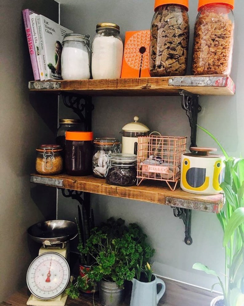 May include: A rustic wooden shelf unit with three shelves, filled with various jars and containers. The top shelf has two glass jars with orange lids, a glass jar with a white lid, and a book titled "Baking". The middle shelf has a variety of jars with different contents, including sugar, tea, and coffee. The bottom shelf has a vintage kitchen scale, a potted plant, and a blue pitcher.