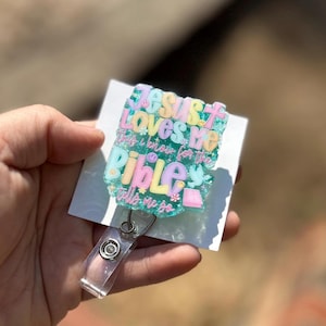 May include: A pastel-colored badge reel with the text "Jesus Loves Me, This I Know for the Bible Tells Me So." The badge reel has a clear plastic clip and a retractable cord. The design includes flowers, a dove, and an open book.