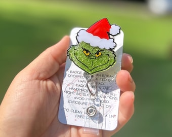 Könnte beinhalten: Ein grünes Glitzer-Badge-Reel mit dem Grinch, der einen Weihnachtsmannhut trägt. Das Badge-Reel ist an einer weißen Karte mit Text befestigt. Das Gesicht des Grinch ist grün mit gelben Augen und einem rot-weißen Weihnachtsmannhut. Das Badge-Reel hat einen transparenten Kunststoffriemen und einen silbernen Clip.