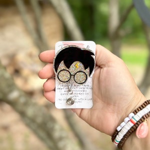 May include: A gold glitter Harry Potter character badge reel with a black and gold lightning bolt design. The badge reel is attached to a silver retractable cord.